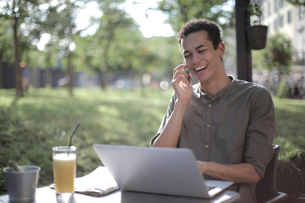 Content African American male freelancer wearing casual clothes sitting at table in street cafe with laptop and speaking on cellphone while working on startup