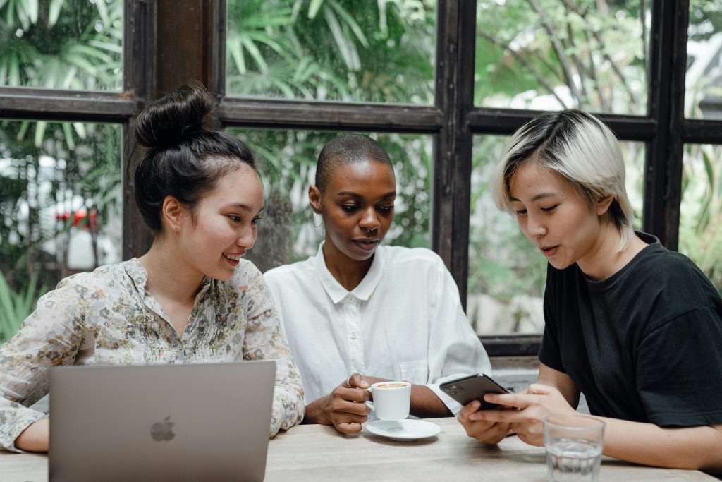 Young Asian and African American women checking posts on social media while sitting in cozy cafe with coffee and laptop near large window with exotic green garden outside